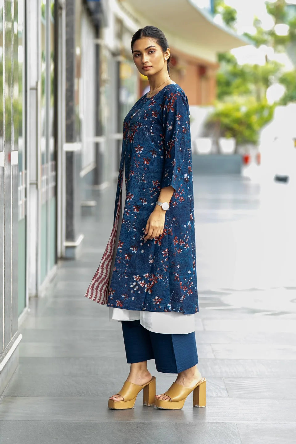 A woman models a navy blue floral kurta with a white underskirt and navy blue wide-leg pants, paired with gold platform sandals, in an outdoor corridor setting.