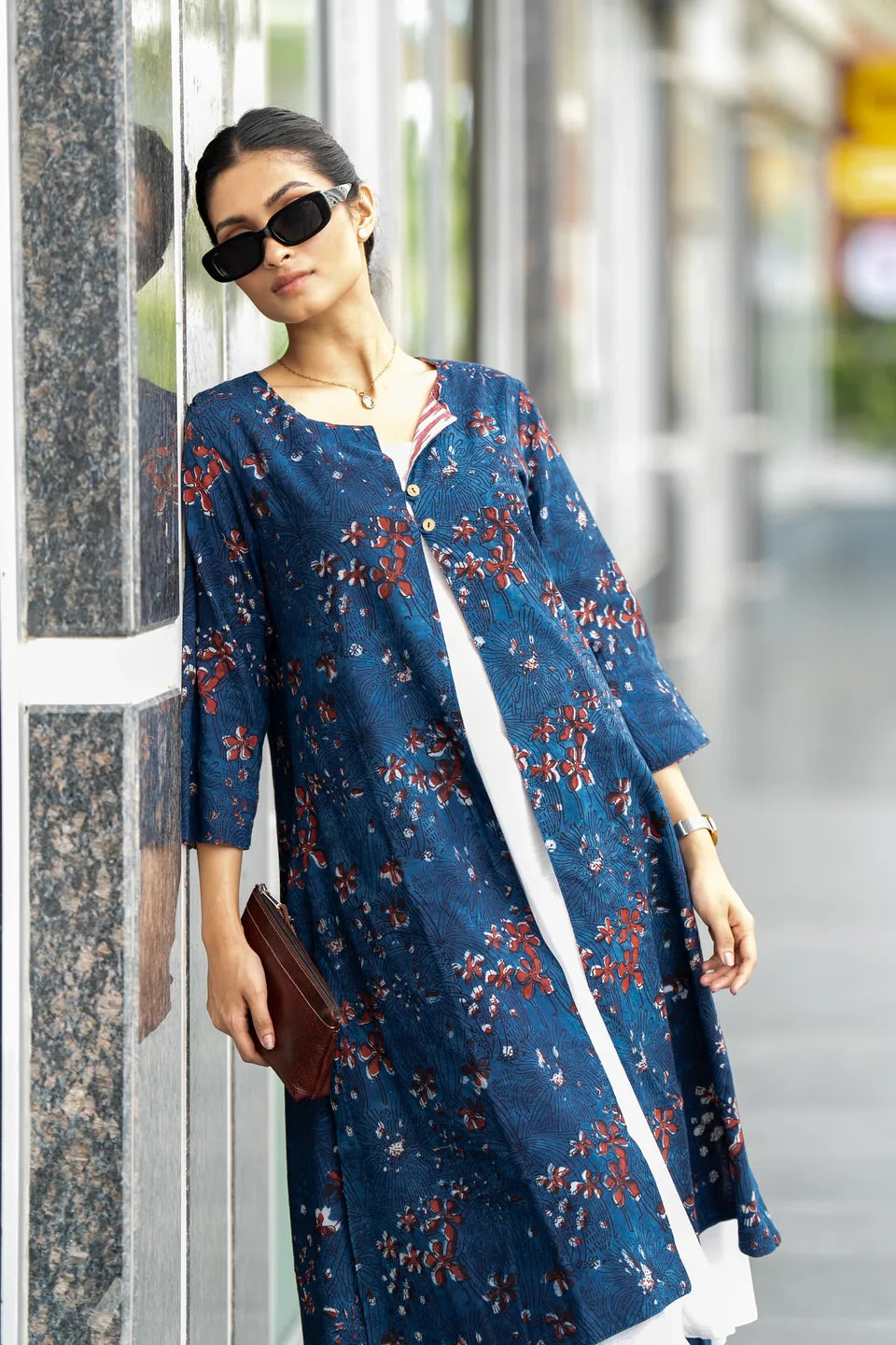 A woman wearing a long blue floral tunic dress with red and orange floral patterns, styled with black sunglasses, a brown clutch, and a white inner dress, standing next to a marble pillar in an outdoor setting.